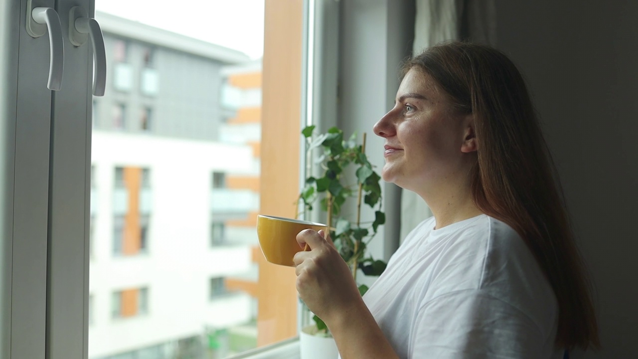 美丽放松的女人，一杯茶或咖啡，坐在家里的窗台上看着窗外视频素材