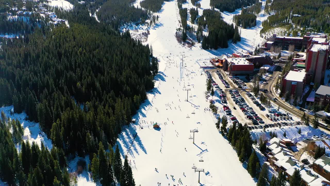 鸟瞰滑雪道，人们乘坐缆车滑下斜坡，旁边是停车场和雪中的松树林。无人机航拍视频素材