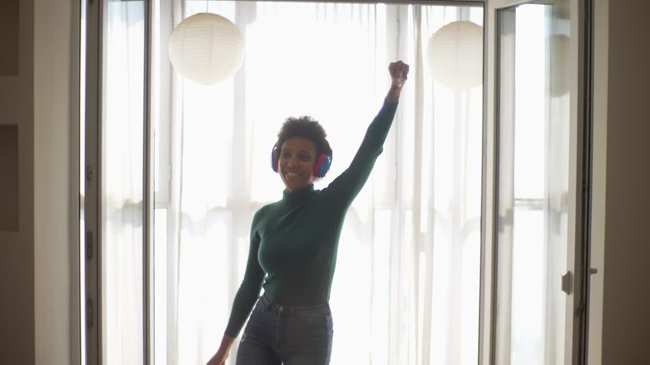 欢乐的年轻非洲裔美国妇女的正面视图，戴着无线耳机，穿着休闲服，在黑暗的客厅靠窗边听音乐跳舞。视频素材