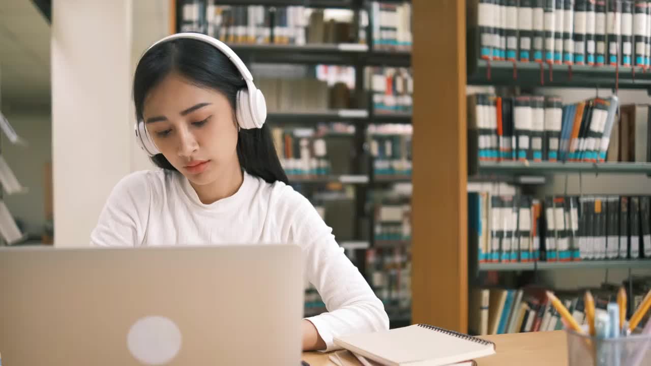 亚洲大学生十几岁的女孩在图书馆里用笔记本电脑读书，为课程做在线研究，为论文作业做笔记，在线教育电子学习概念视频素材