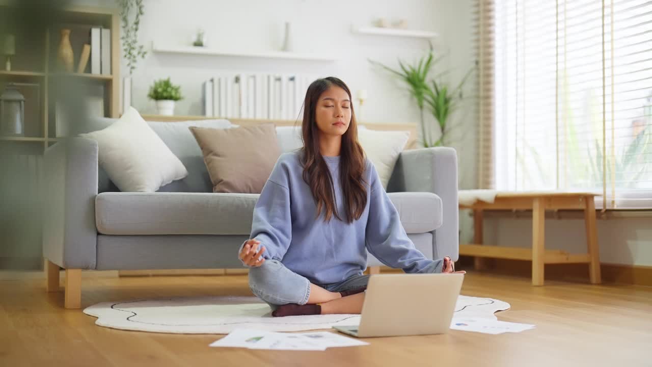 快乐的年轻亚洲女性在家里练习瑜伽和冥想，坐在客厅的地板上，以莲花的姿势，闭上眼睛放松。正念冥想和幸福概念视频素材