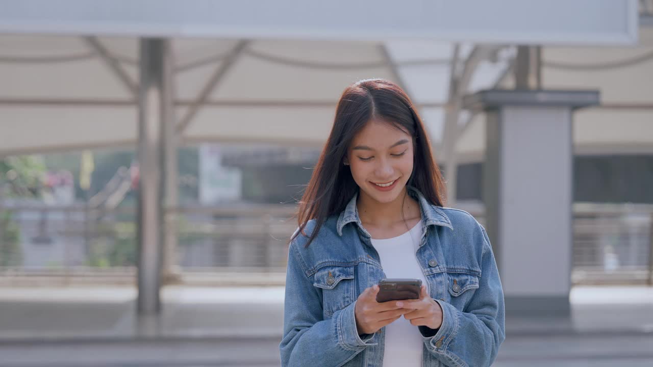 年轻的亚洲女子穿着牛仔裤，夹克，衣服，在晴朗的日子里走在城市的街道上，用智能手机发短信视频素材