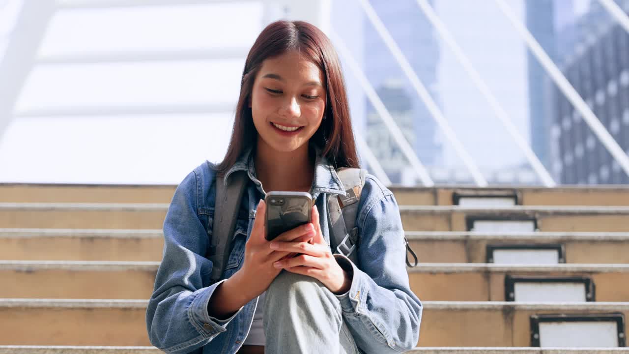 年轻的亚洲女子在户外微笑着使用手机。快乐的女游客穿着牛仔裤夹克，拿着智能手机出现在公共场合视频素材