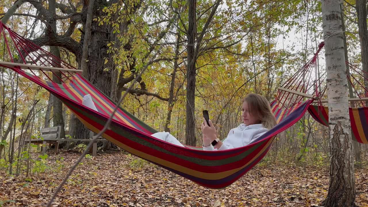 在秋天的森林里，一个穿着浅色运动服的金发女孩躺在摇摆的吊床上，手里拿着一部手机。概念:度假、露营、豪华野营视频素材