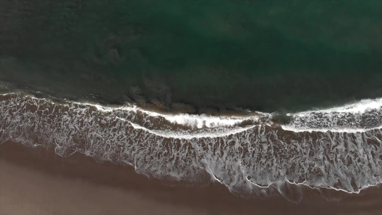 从空中俯瞰的海浪飞溅在海岸线上的沙滩。海浪从上方冲击着空旷的海滩。抽象自然背景旅行。4 k视频素材