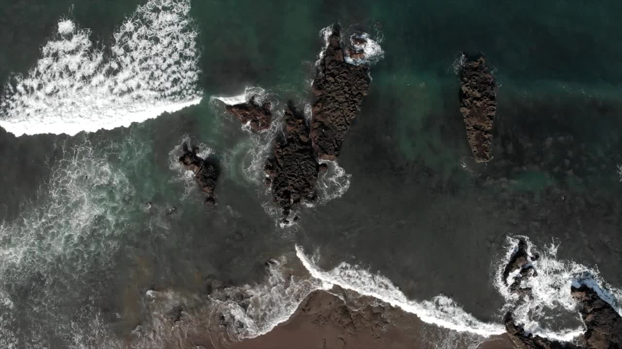 海浪拍打着危险的石头，拍打着空旷的海滩。从空中俯瞰，海浪拍打着海滩附近带有白色泡沫的深色岩石。自顶向下观看无人机4k视频下载