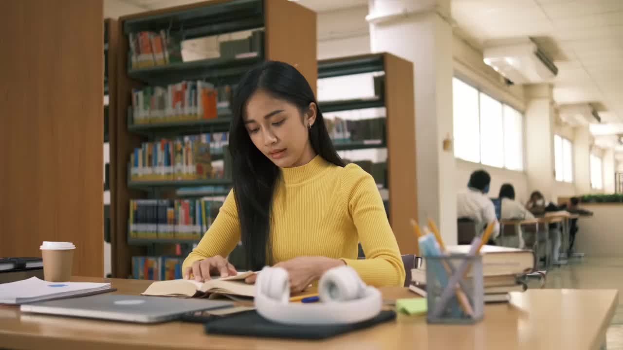 亚洲学生女孩坐在桌边一边看书一边做作业，用笔记本电脑在国外通过网友联系打视频电话，在图书馆里开心地笑着视频素材