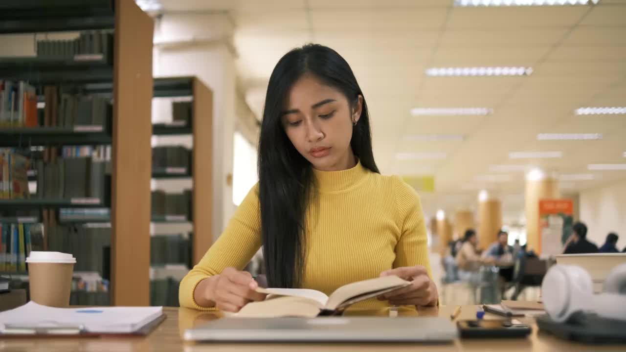 亚洲学生女孩坐在桌边一边看书一边做作业，用笔记本电脑在国外通过网友联系打视频电话，在图书馆里开心地笑着视频素材