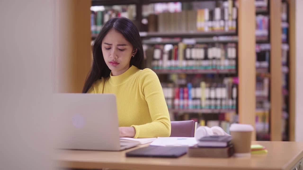 疲惫不快乐压力年轻的亚洲女学生感到疲倦准备考试坐在图书馆桌子在现代大学图书馆过度工作。视频素材