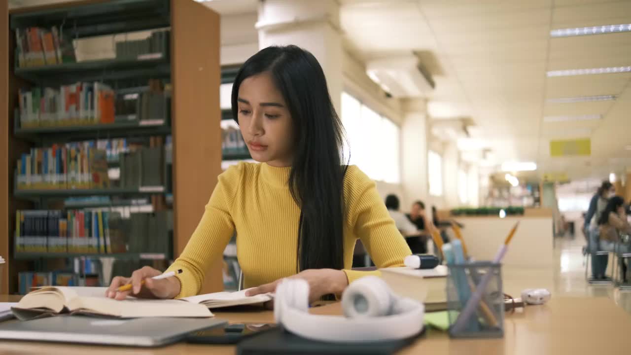 亚洲学生女孩坐在桌边一边看书一边做作业，用笔记本电脑在国外通过网友联系打视频电话，在图书馆里开心地笑着视频素材