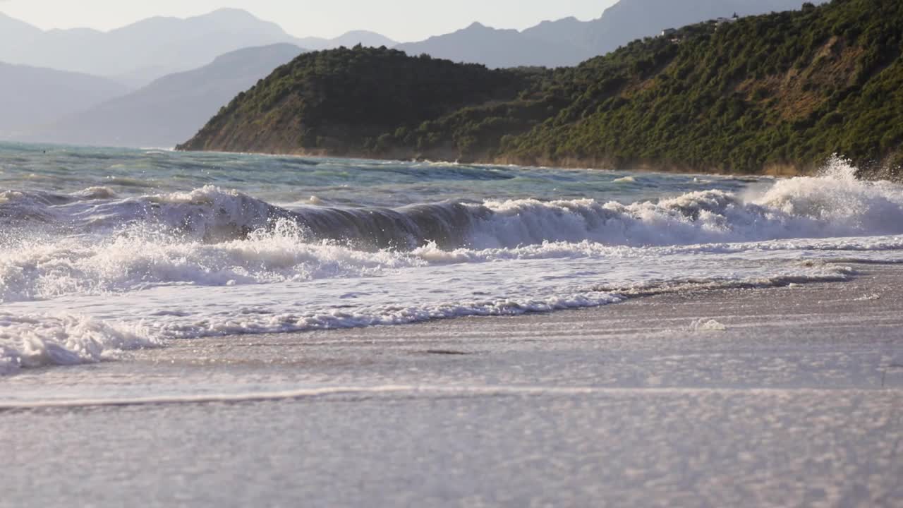 海浪拍打沙滩的慢动作，背景是悬崖和山脉视频素材