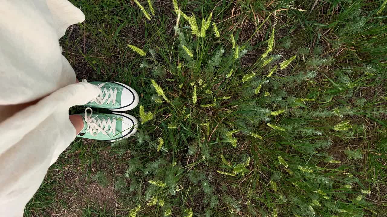 穿着运动鞋和绿草的女性腿的俯视图。草和衣服在风中摇摆。视频下载