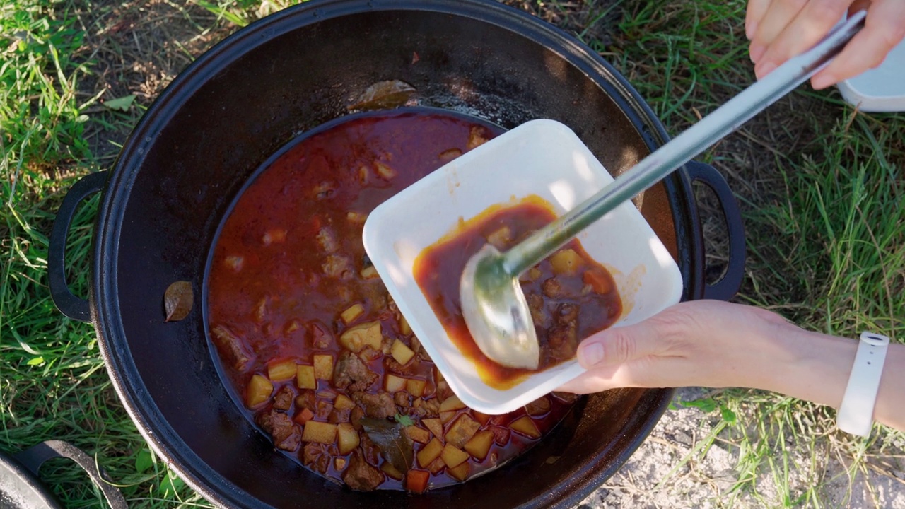 厨师在一次性塑料碗里倒入红辣椒汤，里面有番茄和肉。大锅的特写俯视图，外面放着博格拉奇或罗宋汤，女人的手拿着厨房的勺子。露营的季节。视频下载