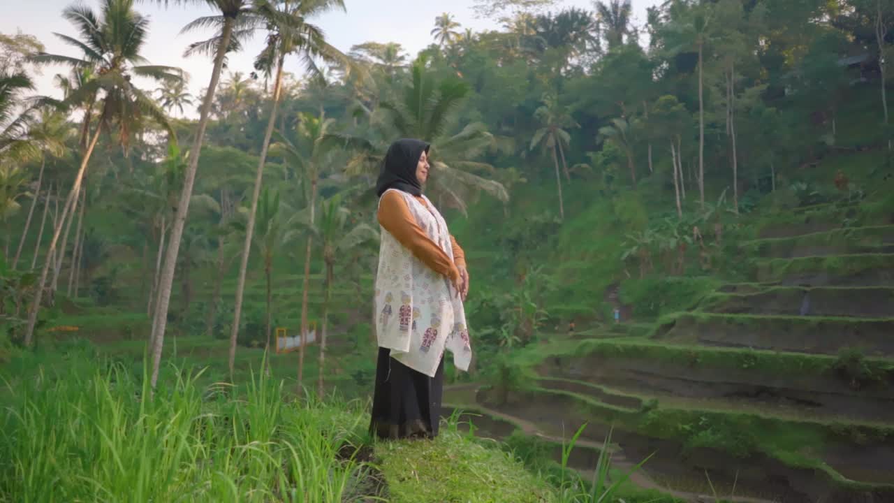 戴着头巾和白色外衣的女性站在稻田里。在早晨欣赏风景。视频素材