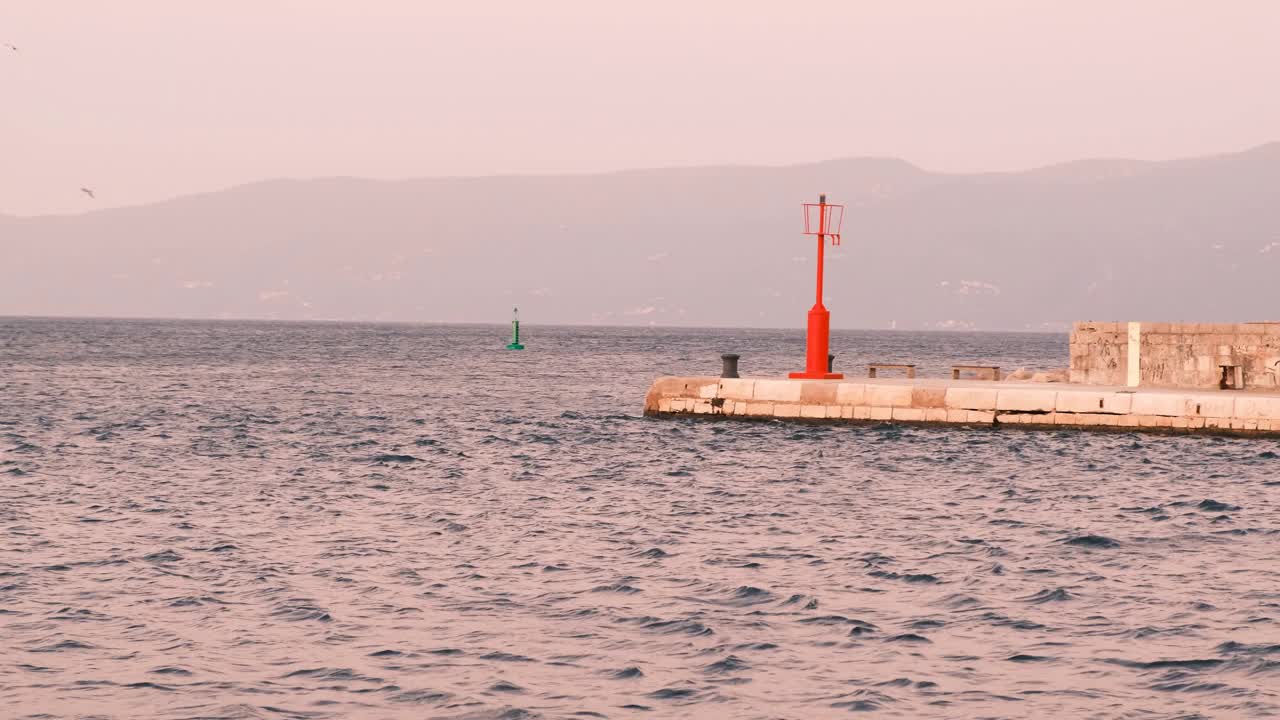 清晨在里耶卡拍摄的静态海景海景，港口码头上的红色灯塔，飞翔的海鸥和被海浪移动的绿色导航安全浮标。视频下载