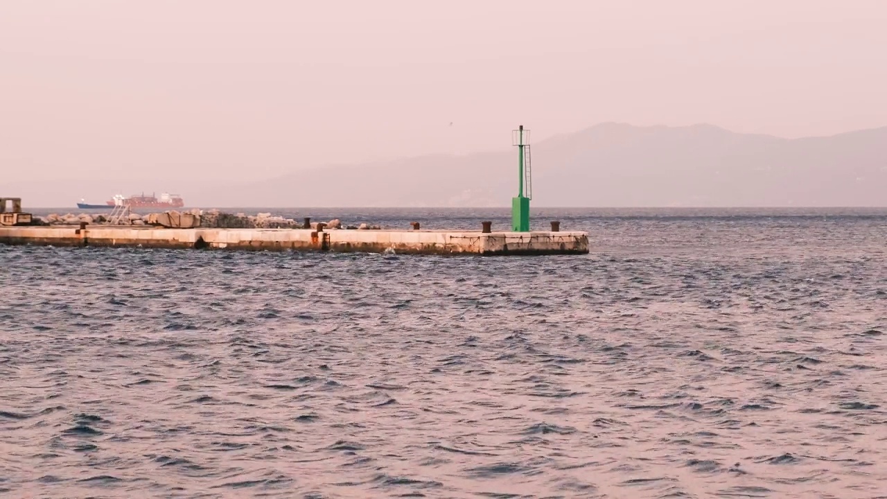 清晨在里耶卡拍摄的静态海景海景，背景是港口码头上的绿色灯塔和一艘集装箱船。由于刮风天气，海面上泛起了涟漪。视频下载