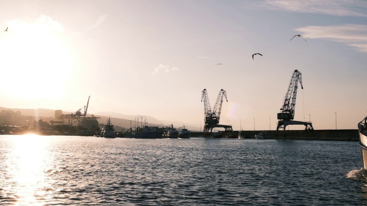 里耶卡港口的阳光明媚的早晨，过去的老起重机的剪影点缀着全景，背景是摩天大楼。一艘渔船在茫茫大海上的场景视频下载