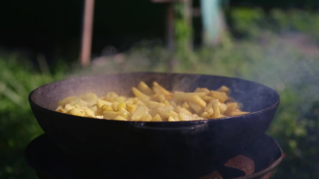 在室外用煎锅煎土豆。在特制的铸铁盘子上煎土豆片。在明火上烹饪简单的食物，是户外休闲或野餐的概念。4K视频片段。视频下载