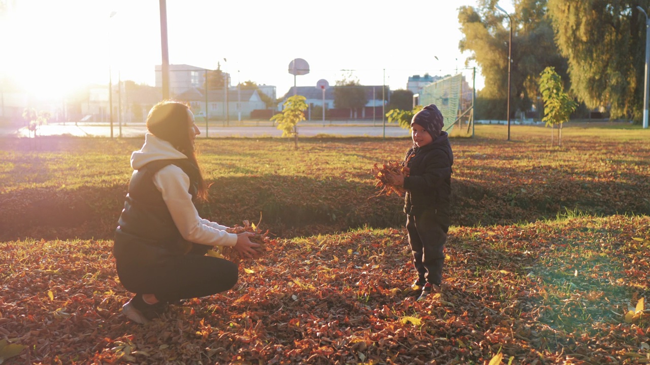妈妈和小男孩在头顶上扔了一堆树叶，看着它们飘落。孩子在秋天的公园里玩金色的树叶。幸福的家庭在秋天散步。公园里的人。幸福家庭概念。视频素材