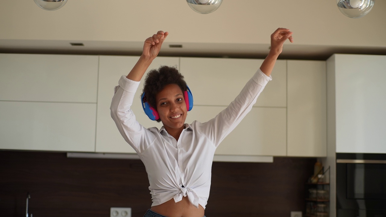 无忧无虑的年轻混血女性在家里跳舞，戴着耳机听音乐，在现代公寓室内举起手臂。视频素材
