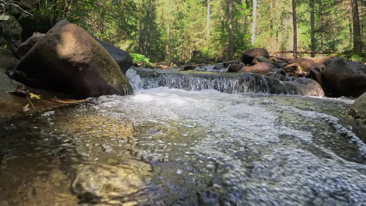 相机缓缓跳入高山河流中最纯净的充满气泡的水中。半水下慢动作拍摄的河流在绿色的森林和山脉。石头之间有小瀑布的小溪视频素材