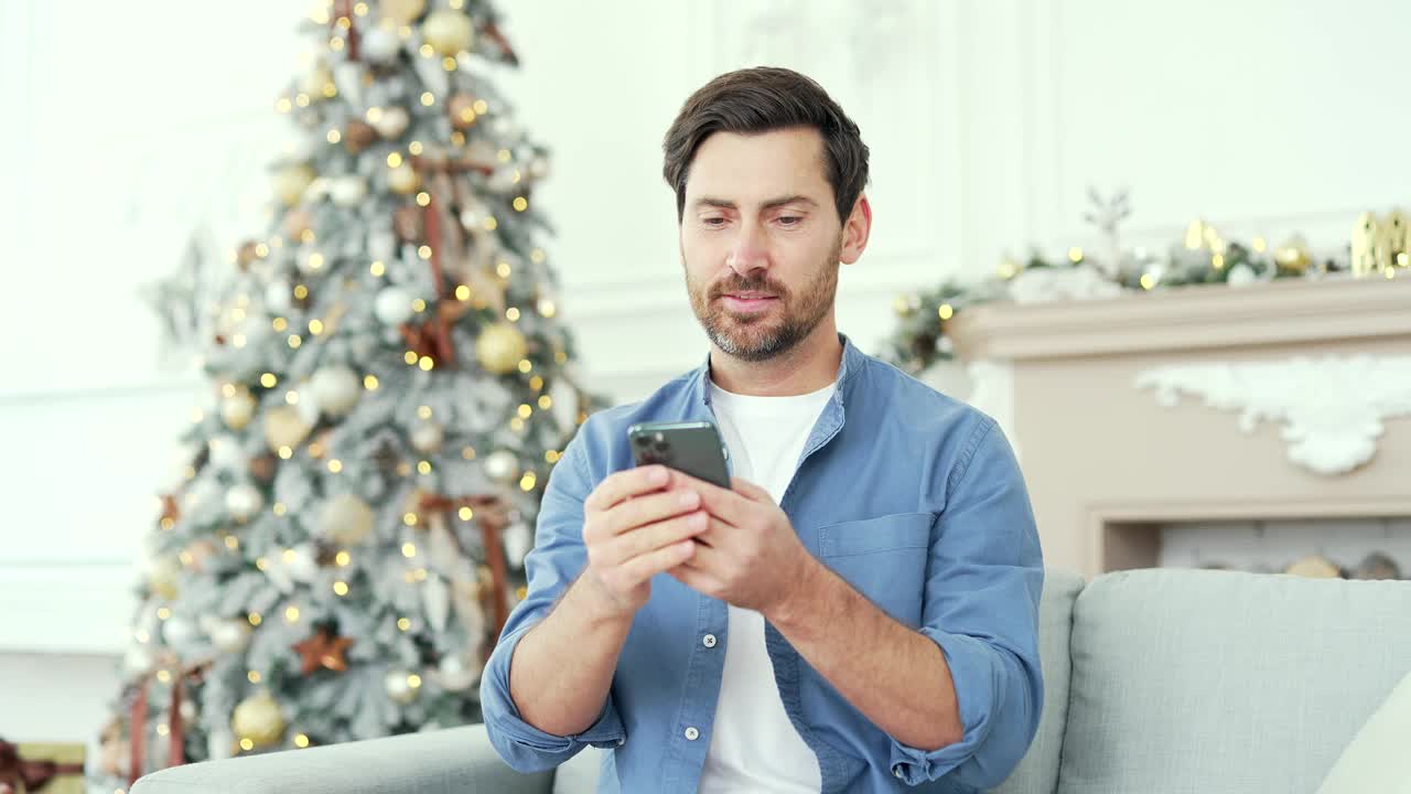 冬季新年圣诞假期，一位满脸笑容的大胡子男子坐在家里的沙发上玩智能手机视频素材