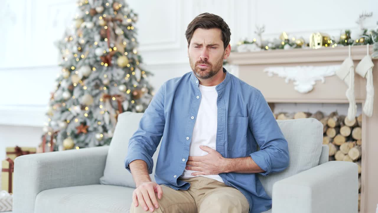 一名心烦意乱的男子在冬季新年圣诞假期坐在家里客厅的沙发上感到胃痛视频素材