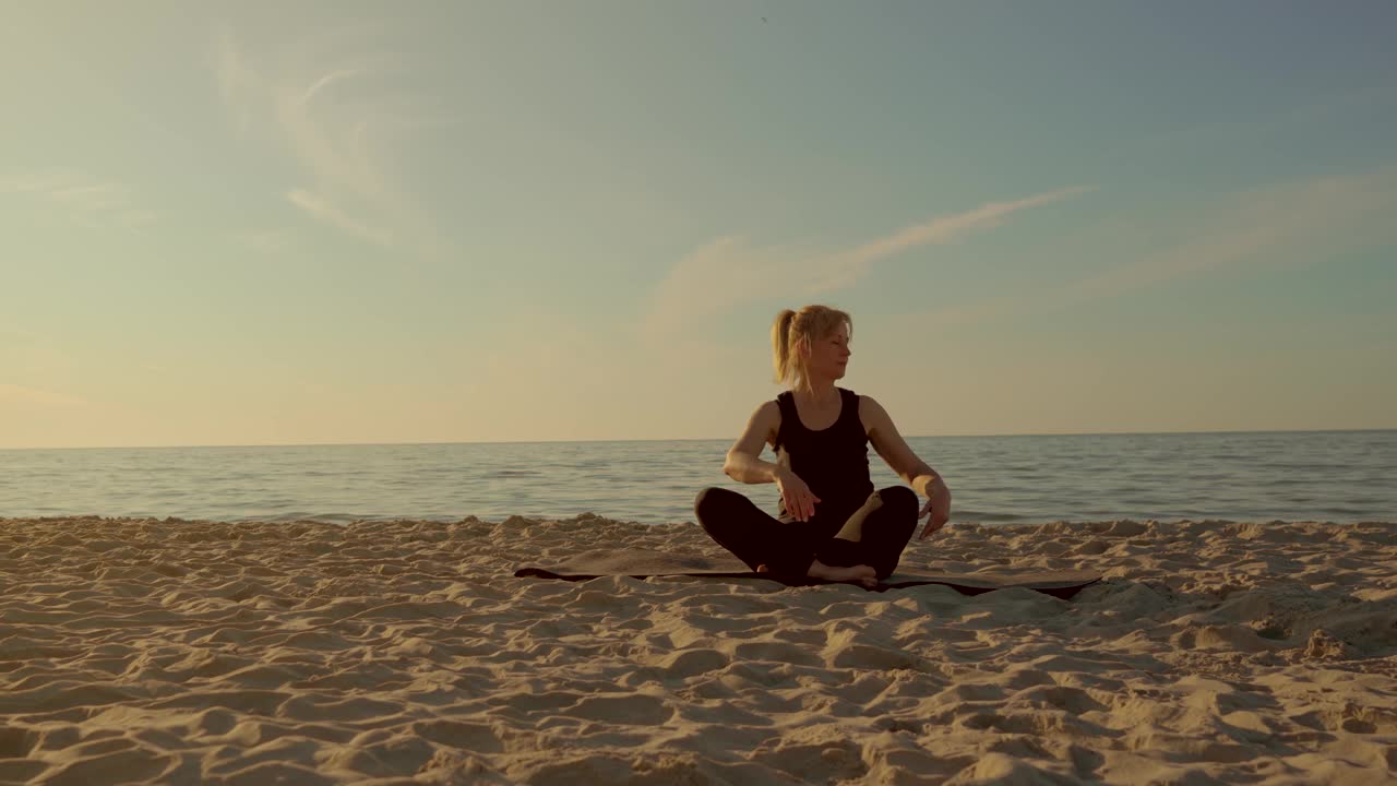 夏夜，一名专注的女子在沙滩上练习冥想。坐在海浪边的健身垫上做瑜伽的女人视频素材