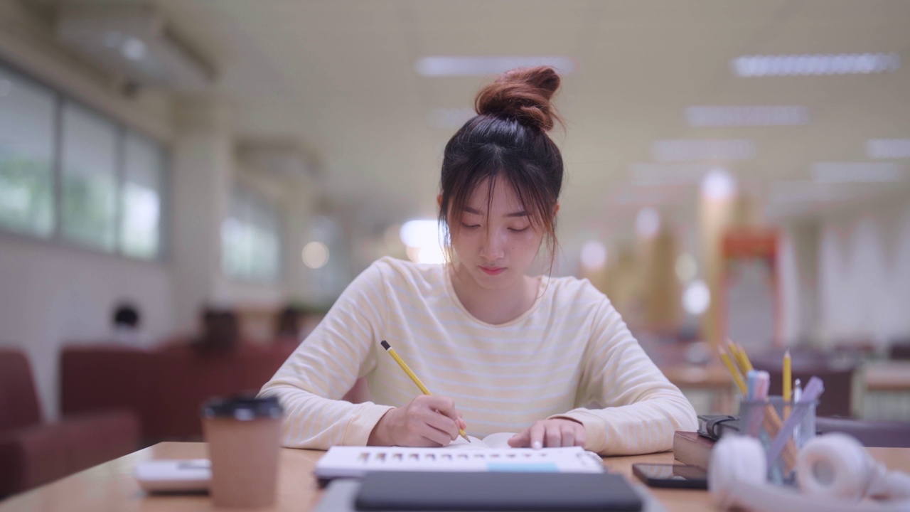 一个勤奋的亚洲女学生在学校或大学图书馆里忙着总结她所学的课程或准备考试。她专注于她的工作，利用安静的空间视频素材