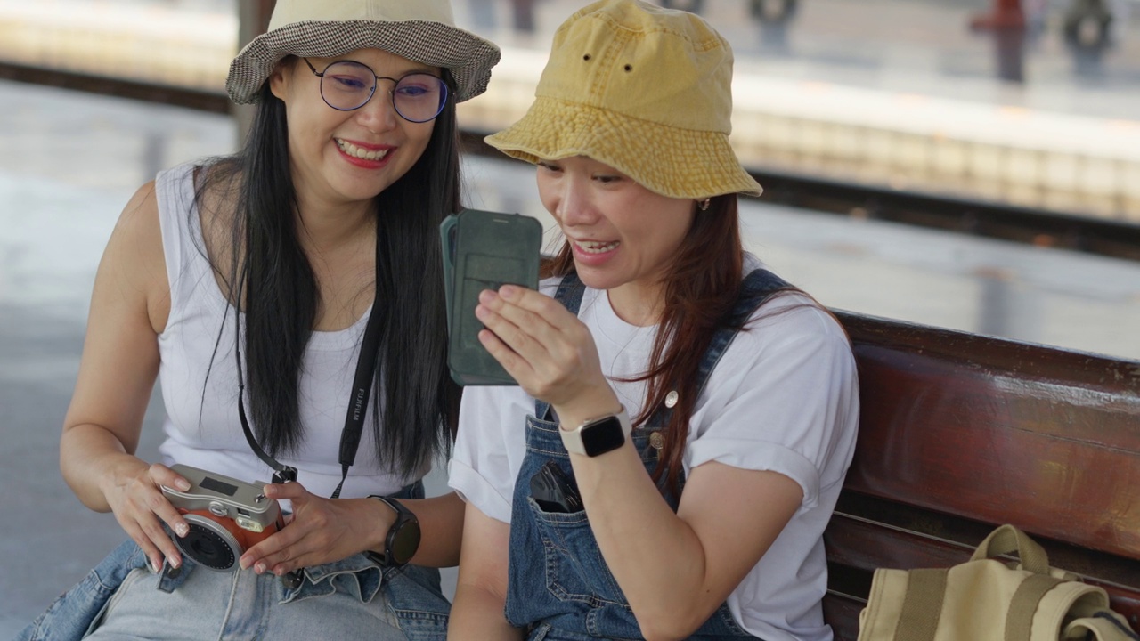 两个快乐的亚洲旅行者朋友在火车站等火车时用手机通过社交媒体网站与朋友联系视频素材