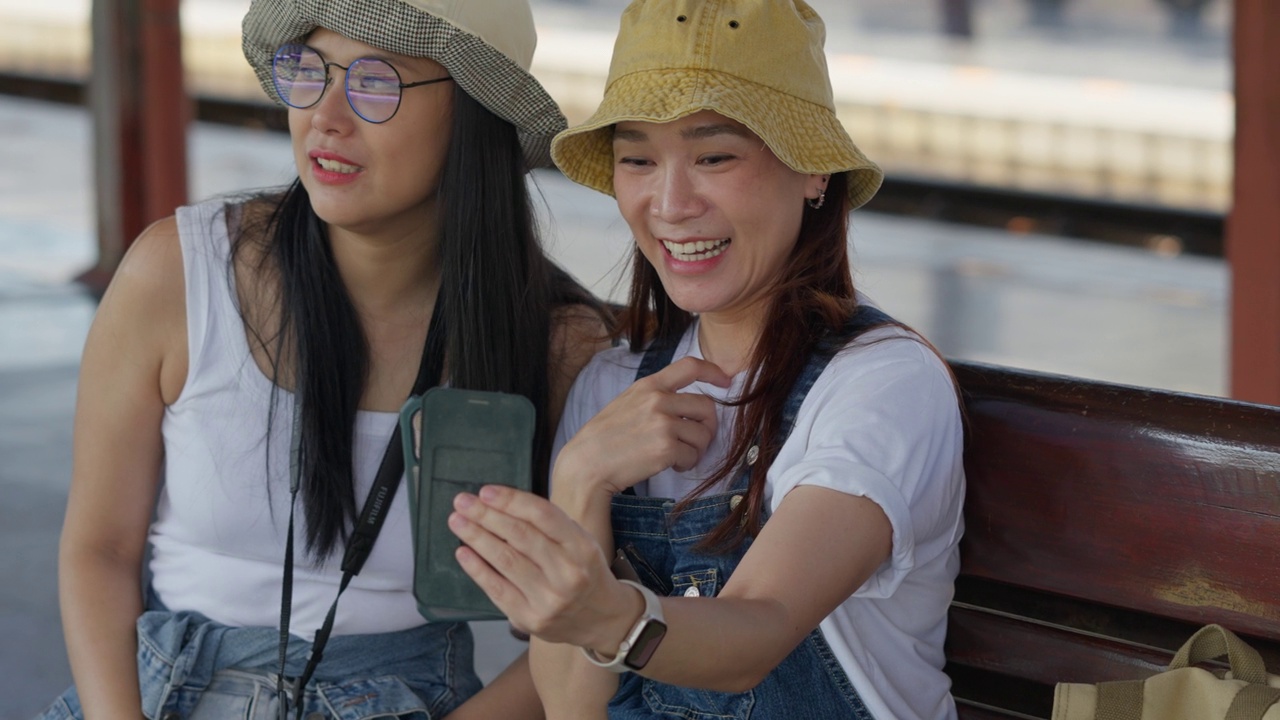 两个快乐的亚洲旅行者朋友在火车站等火车时用手机通过社交媒体网站与朋友联系视频素材