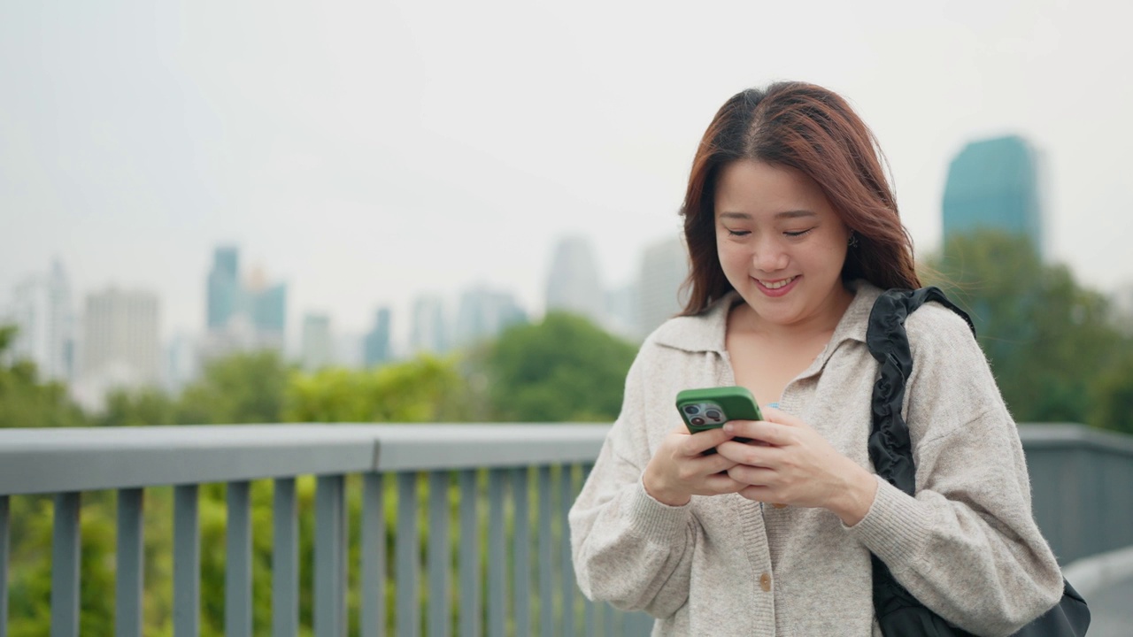 微笑的年轻亚洲女子手持智能手机走在街上。美女边走边用手机，市景。科技与生活理念视频素材