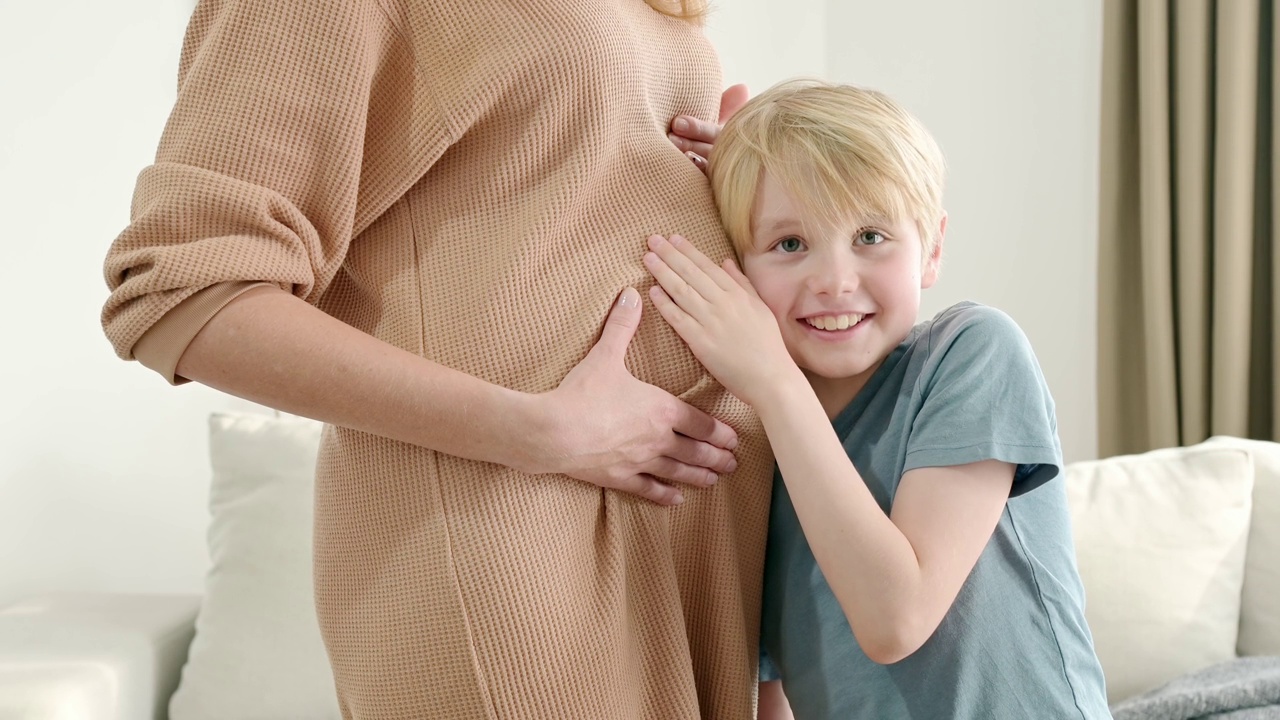 一个男生抱着他怀孕的妈妈，倾听着她的肚子。怀孕的母亲和她的大孩子在家里。未来的大哥。视频素材