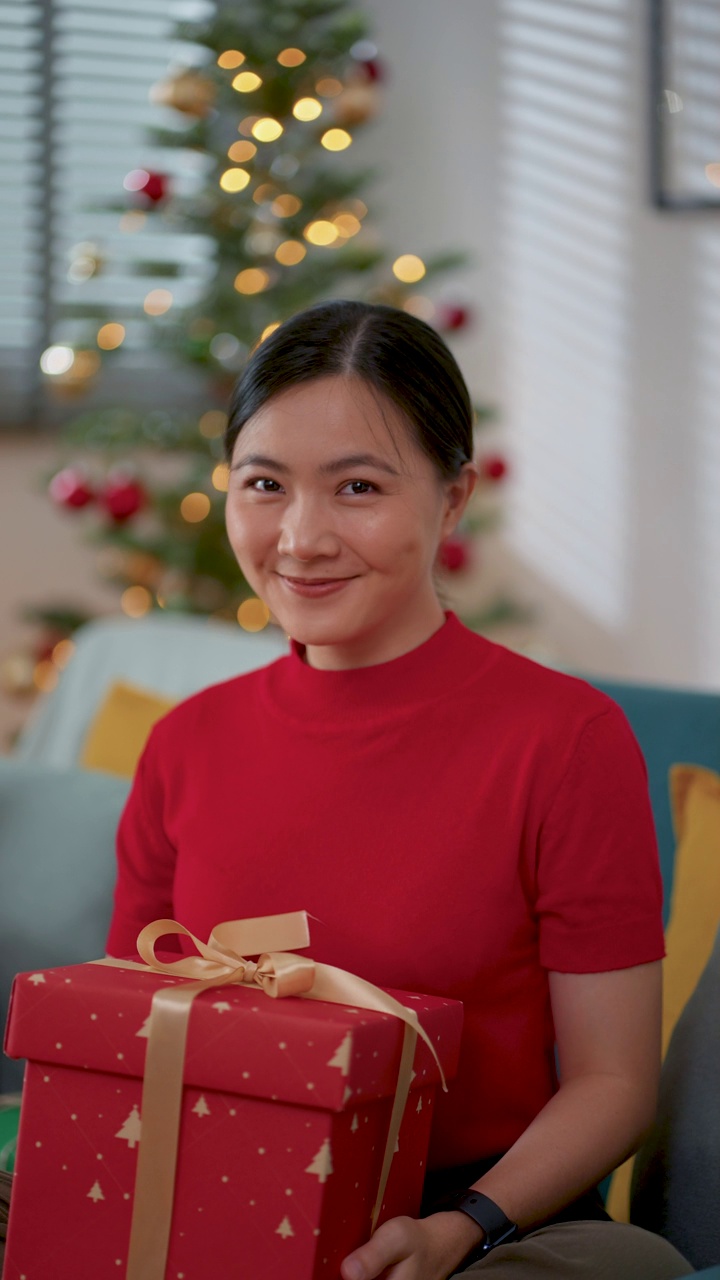 圣诞节那天，坐在客厅里的一名亚洲女子开心地微笑着向镜头展示了一个礼品盒。视频素材