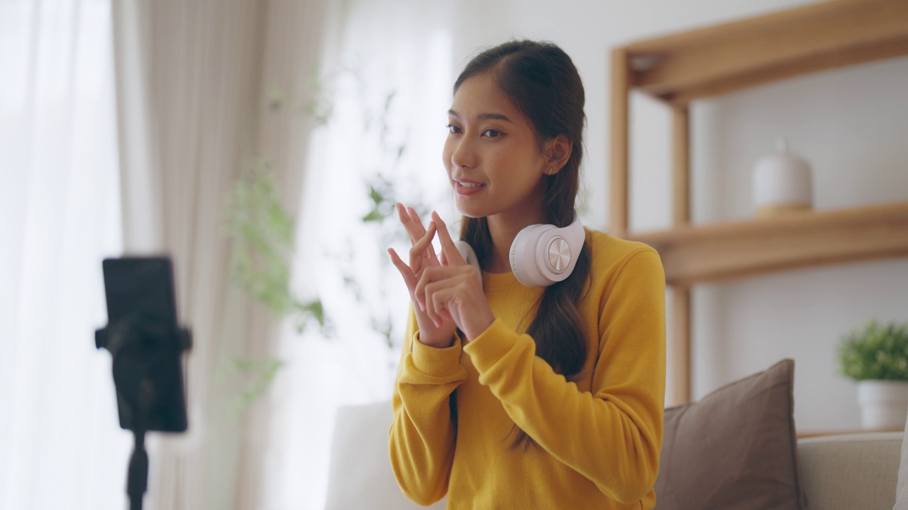 快乐的年轻亚洲女人在家里放松，微笑的女人坐在沙发上，看着手机屏幕上的手机，女孩用视频通话给朋友视频素材