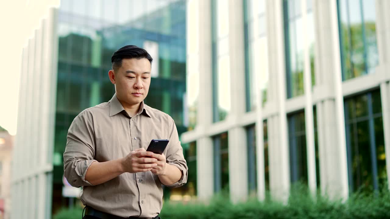 一位自信的商人穿着衬衫，用智能手机走在办公楼附近的街道上。视频素材