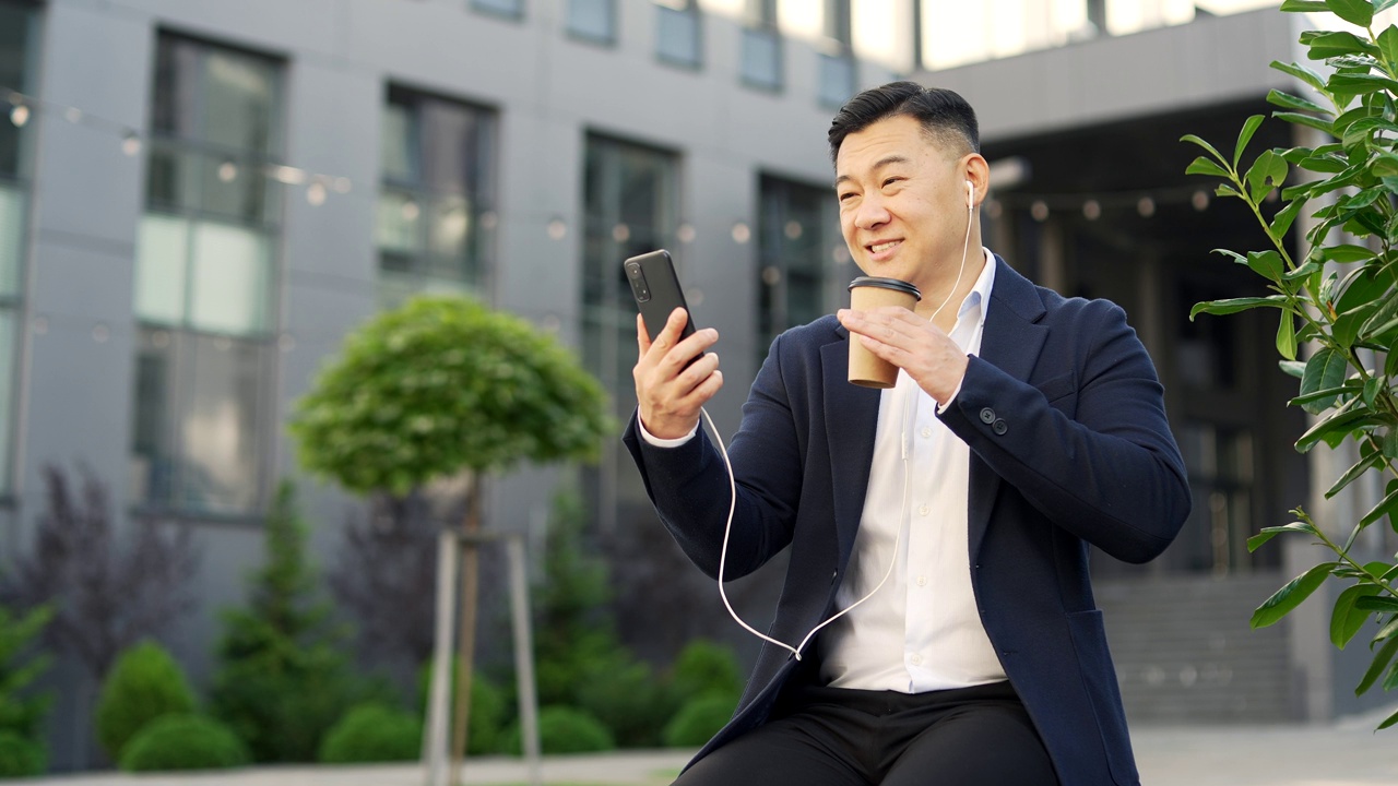 一名身着正装的亚洲商人坐在办公大楼附近街道的长椅上用智能手机进行视频通话。视频素材