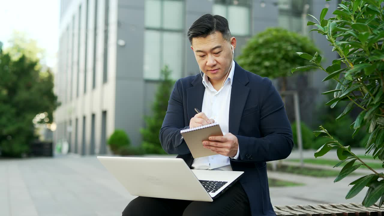 一名身着正装的亚洲商人坐在办公大楼附近街道的长椅上，用笔记本电脑做笔记，正在进行视频通话。视频素材