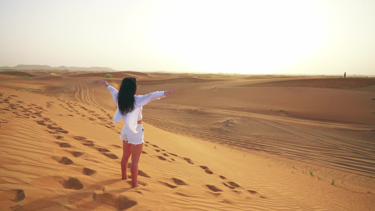 后景年轻美丽的白人妇女站在沙漠和举起她的手。在炎热的迪拜旅游。观光路线。感受暑假的快乐和自由。视频素材