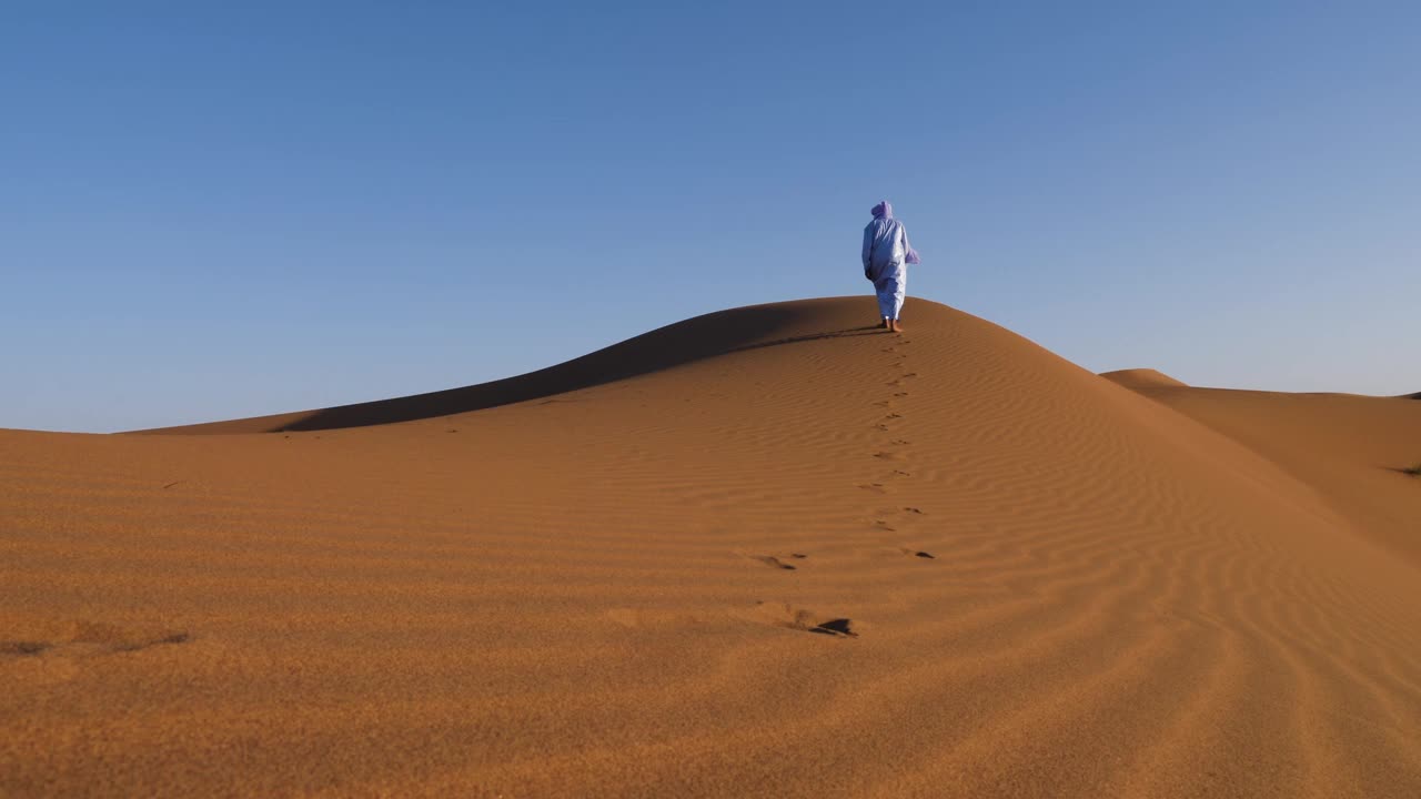 摩洛哥，埃尔格切加加沙漠，一名穿着传统服装的摩洛哥男子身穿蓝色gandoura，长袍和头巾走在沙丘上。视频下载