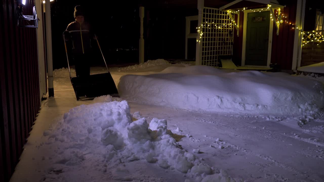 冬天，下班后的晚上，一名男子在房子附近的小路上清理积雪。一个男人用铁铲铲雪。视频下载