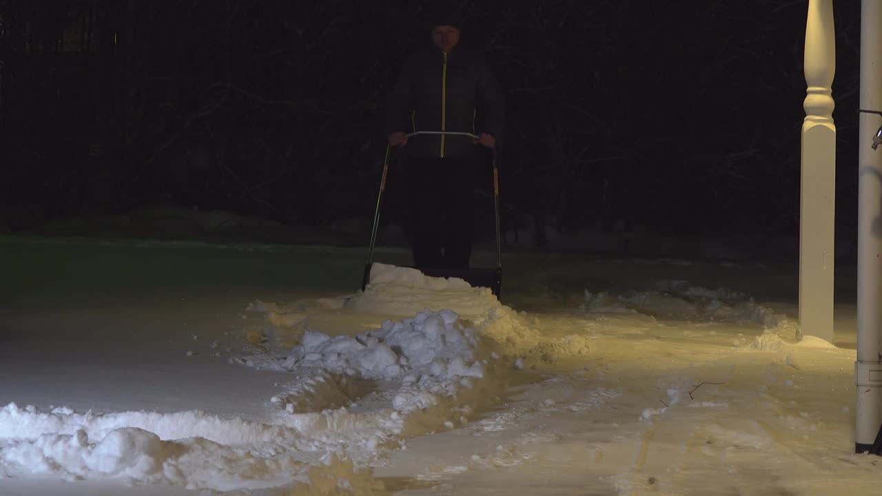 冬天晚上下班后，一名男子在车道上清扫积雪。一个男人用铁铲铲雪。视频素材