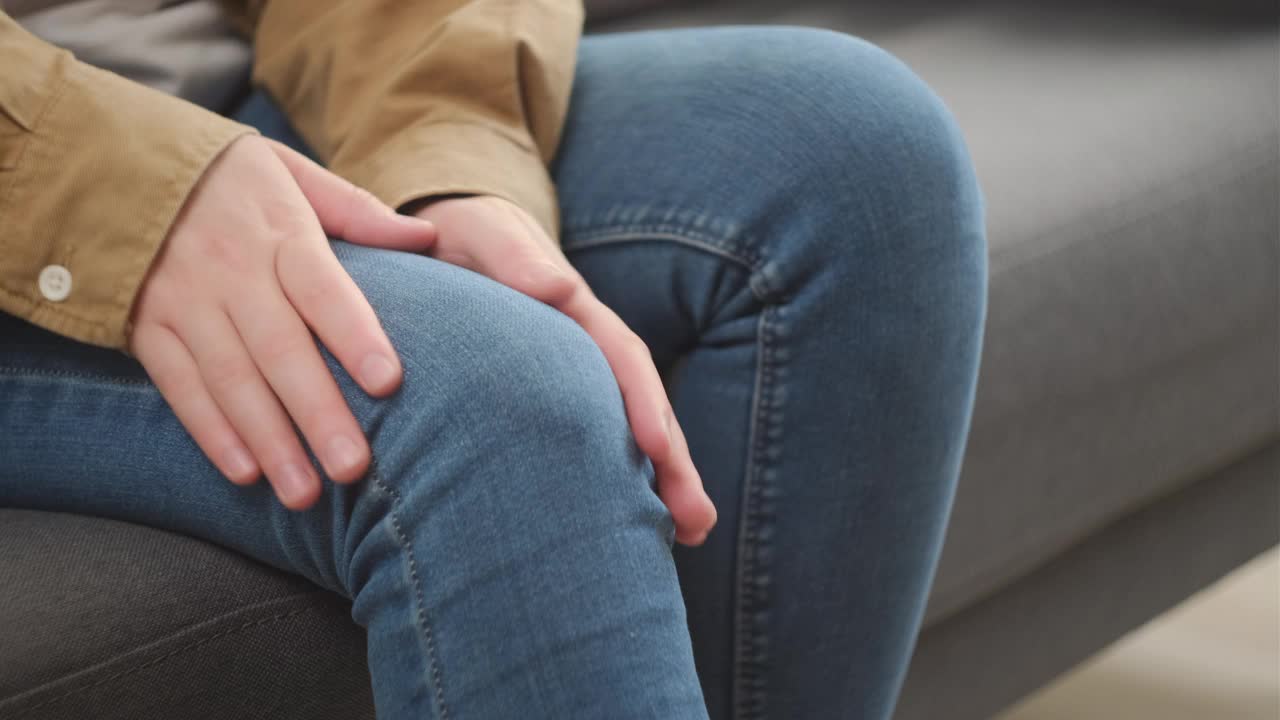 女性坐在家里沙发上按摩膝盖腿部肌肉的特写。不知名的年轻女子患有严重的膝关节疼痛，不能走路，疾病，痉挛，风湿病概念视频素材