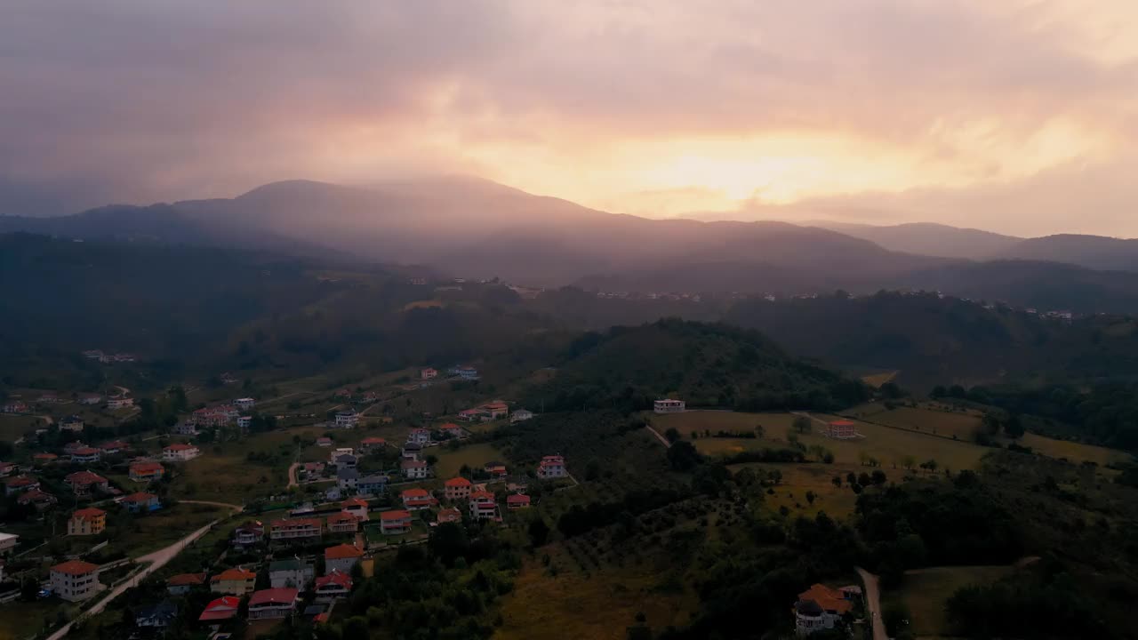 高空无人机拍摄的山腰村庄，背景是多云的黄昏天空下的山脉视频素材