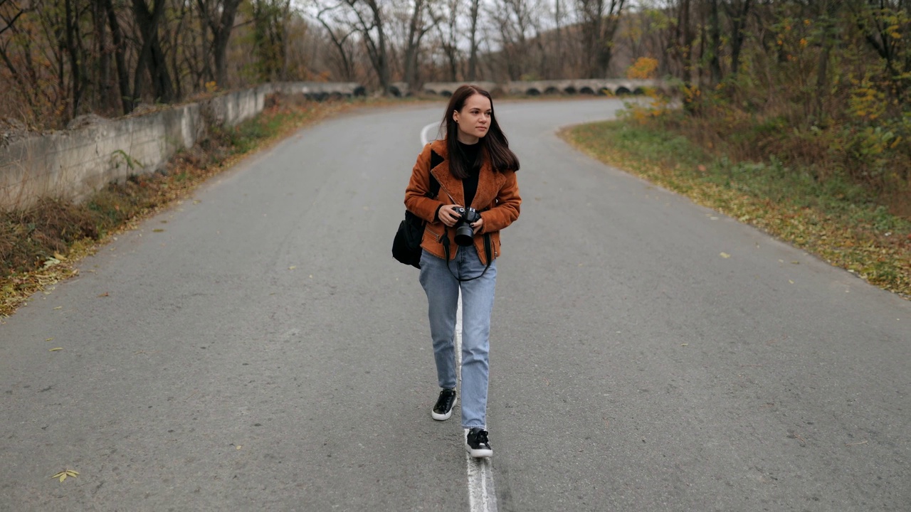 年轻迷人的女人背着背包和相机走在美丽的道路上，穿过森林，独自在户外旅行，享受假期视频素材