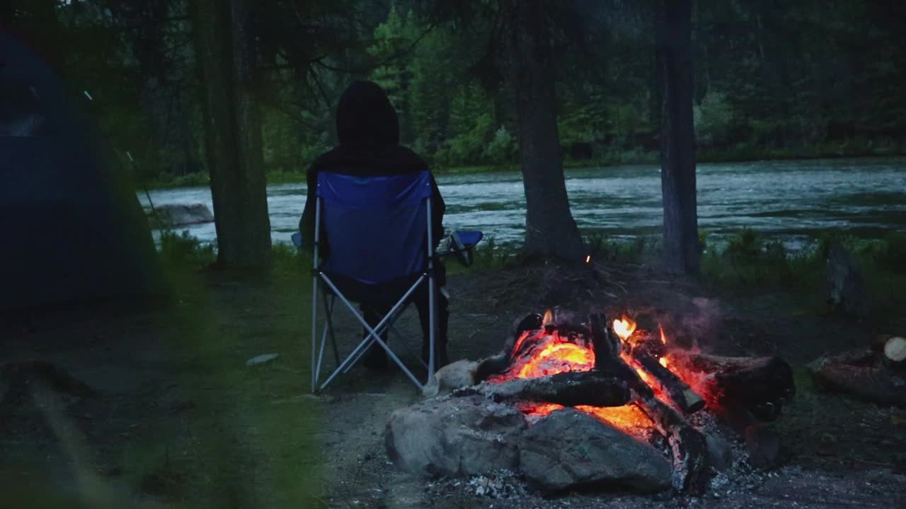 自由旅行者在山间河边靠近火场的地方远程工作。晚上坐在露营地的椅子上视频下载