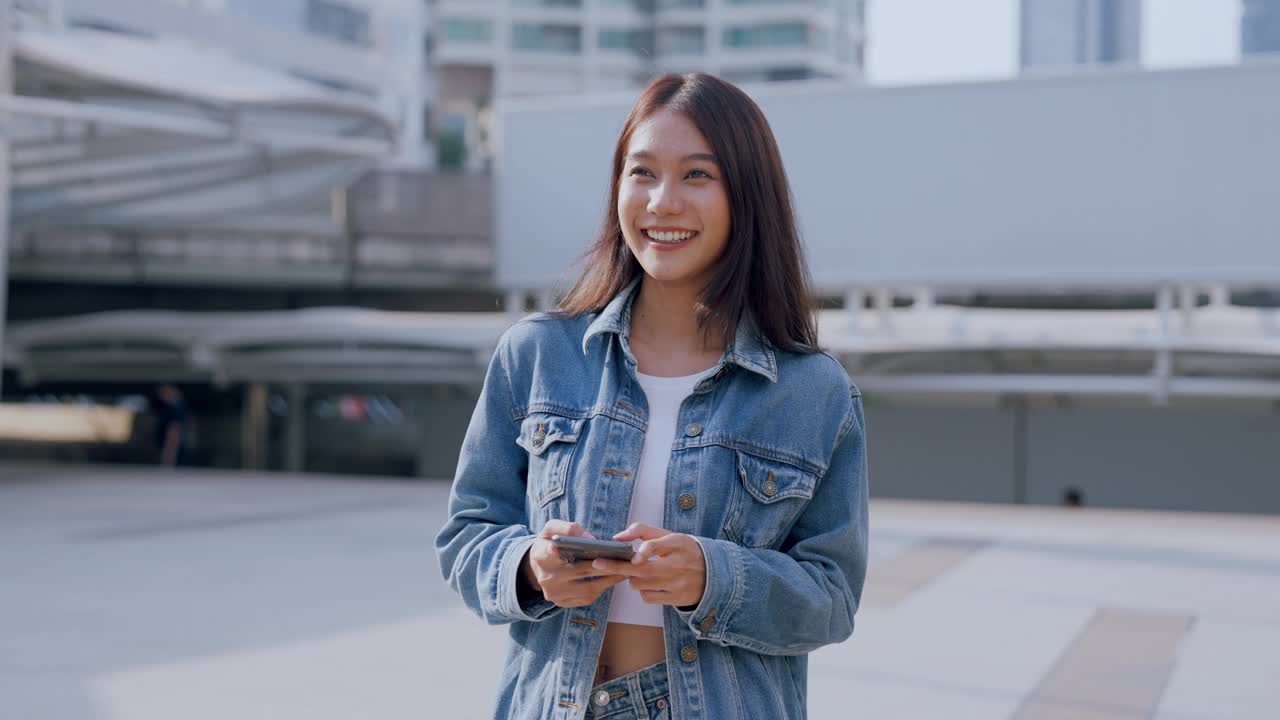 年轻的亚洲女子穿着牛仔裤，夹克，衣服，在晴朗的日子里走在城市的街道上，用智能手机发短信视频素材