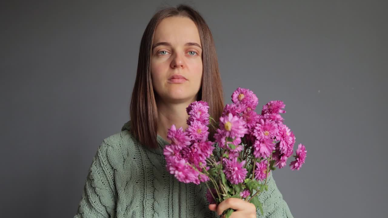 花粉的季节。鼻炎反应。菊花过敏。季节性疾病。生病的女人穿着绿色针织衬衫，手里拿着花束，孤立在灰色的背景下，脖子发痒视频素材