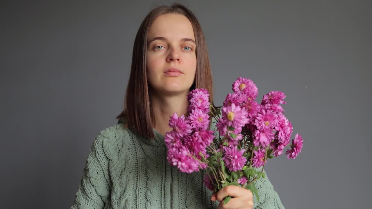 生病的不高兴的女人穿着绿色针织衬衫，手里拿着花束，闻着菊花的气味，打喷嚏，在灰色的背景上遭受过敏。视频素材