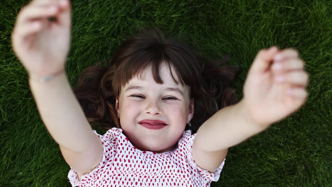 国际儿童节。快乐的小女孩躺在绿色的草地上，看着，伸出她的手，对着镜头挥手。微笑的孩子在户外的草坪上看着天空，玩得很开心，做着梦。视频素材